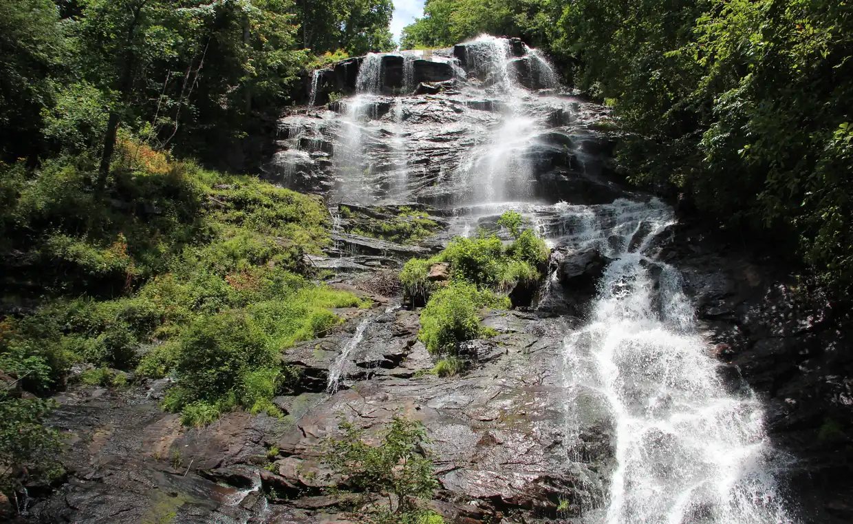 A tall, stepped waterfall streams down dark rock between green trees and ferns in bright summer light.