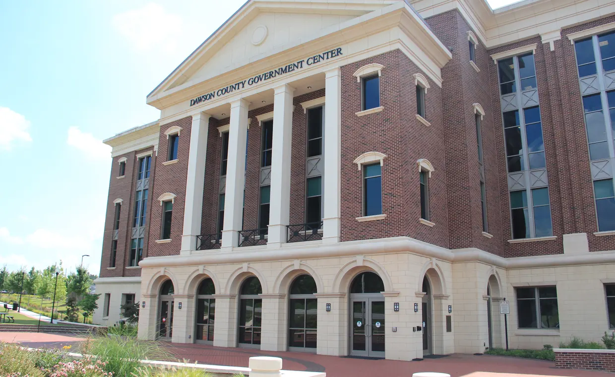 A red brick government building with cream trim and a columned, pedimented entrance carries the lettering Dawson County Government Center; an arched ground-floor arcade, a brick plaza and young landscaping fill the foreground under a blue sky.