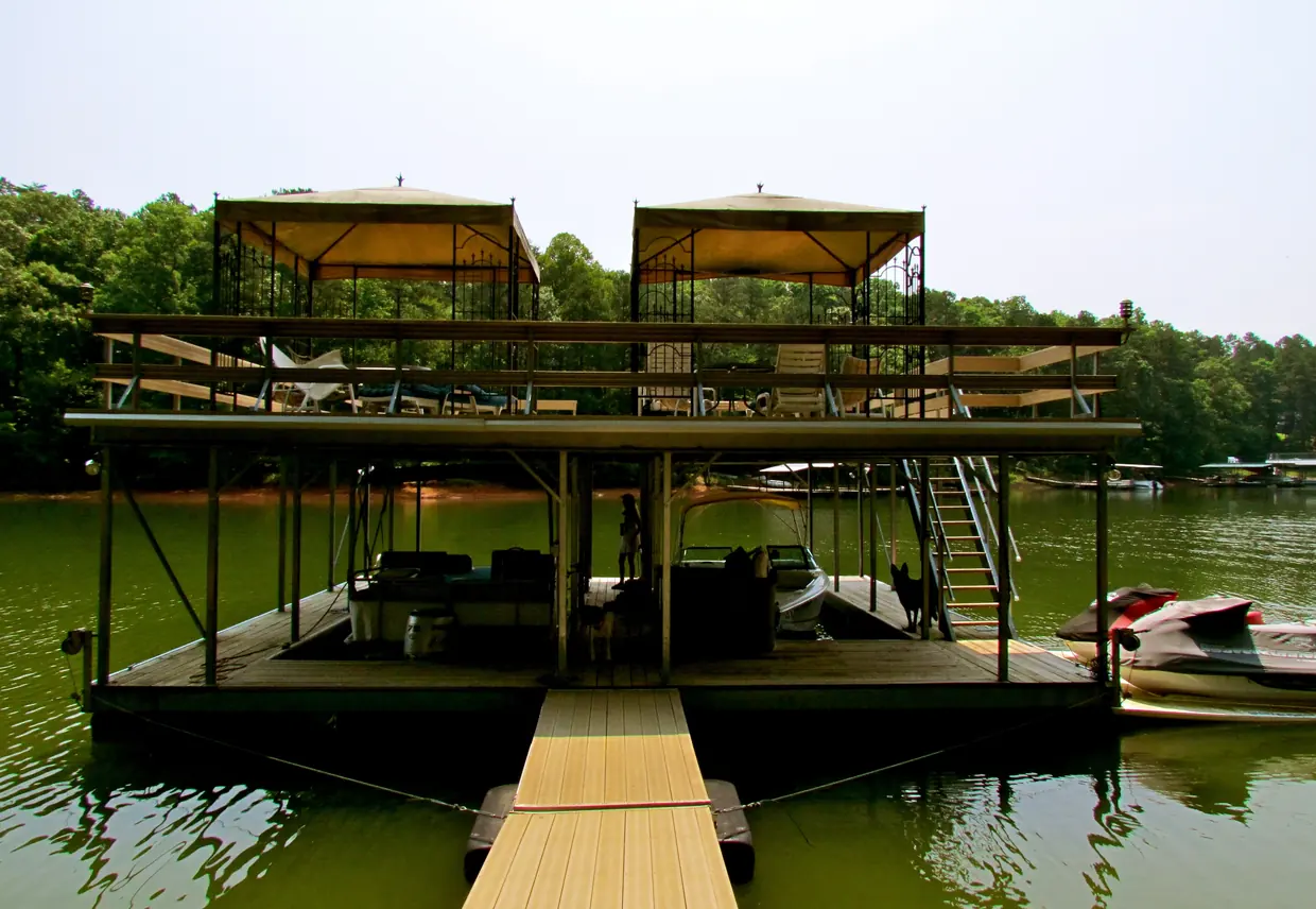 A wooden walkway leads out to a large two-level floating dock: the shaded lower level holds a pontoon boat and a bowrider in their slips, the upper sun deck has railings, lounge chairs and two square fabric canopies, and a personal watercraft is tied alongside; green water and wooded shoreline with other docks fill the background under a hazy white sky.