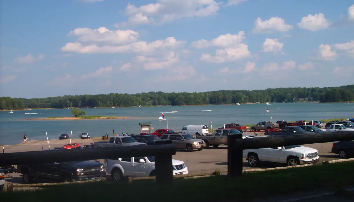 Beyond a dark wooden rail fence and a parking lot full of pickup trucks and SUVs with boat trailers, a boat ramp slopes into a broad blue lake dotted with motorboats, a small sailboat and a low grassy islet with one tree, backed by a continuous wooded shoreline under a blue sky with scattered white clouds.