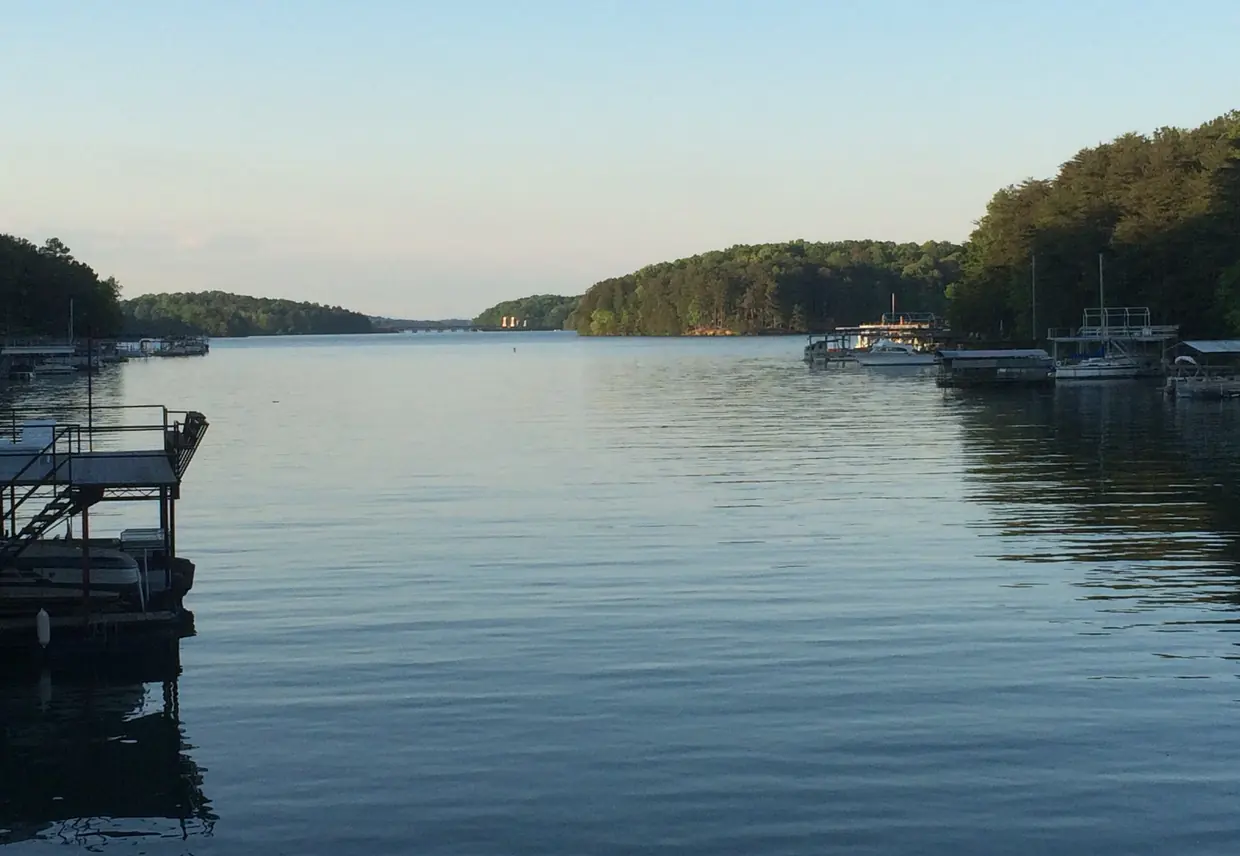 Calm, glassy water fills a long cove that narrows toward a distant gap; on the right a row of covered boat docks with white boats lines a steep wooded bank, on the left a dark metal dock with stairs stands in the foreground, and the far wooded hills glow in low evening light under a clear pale sky.