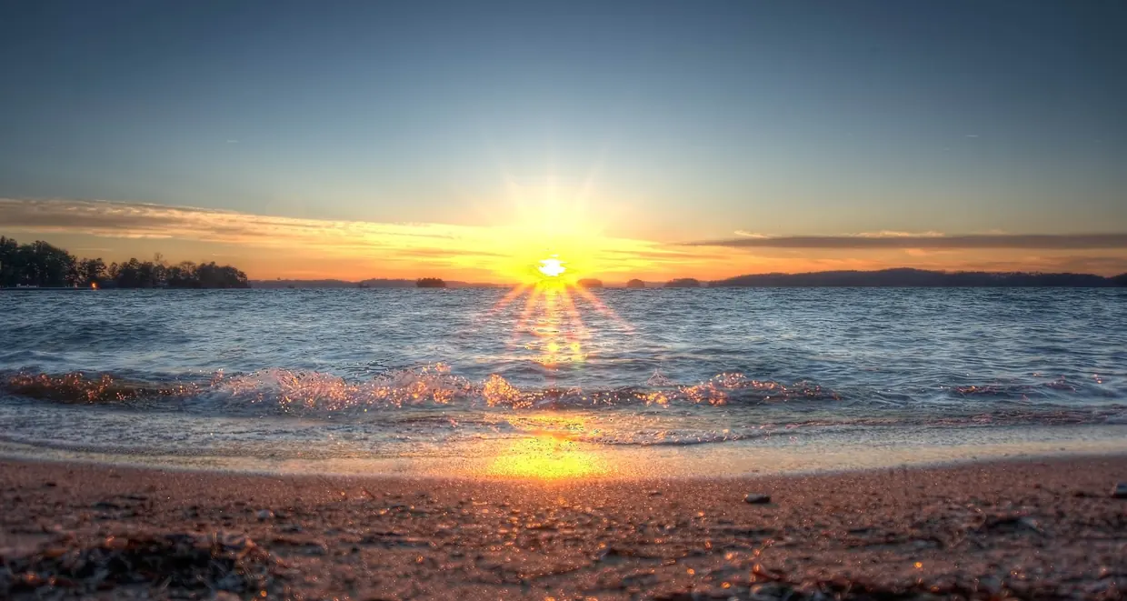 The sun sits on the horizon in a starburst over wide, gently choppy water, laying a golden path to a small breaking wave and a wet sand beach in the foreground; low wooded islands and points line the horizon under a clear sky fading from gold to deep blue.