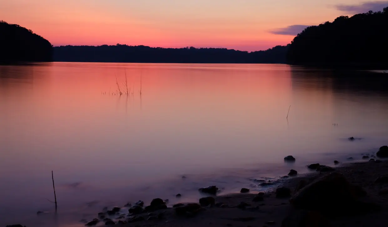 A long exposure at dusk turns glassy lake water pink and violet beneath an orange-to-pink sky; dark wooded hills frame a cove on both sides, a few dead stems poke from the water, and smooth dark stones dot a sandy beach in the foreground.
