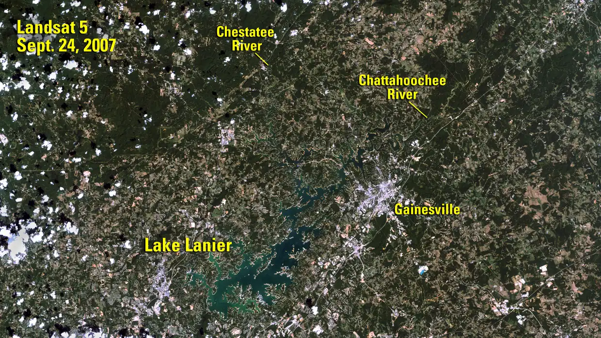 A natural-colour satellite view of north Georgia countryside, partly flecked with small white clouds, shows a thin, pale blue-green branching lake with light tan edges; yellow labels read 'Landsat 5, Sept. 24, 2007', 'Lake Lanier', 'Chestatee River', 'Chattahoochee River' and 'Gainesville'.