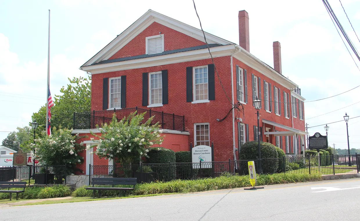 A two-story red brick building with a gabled front, white-framed windows with dark shutters and two chimneys stands behind a black iron fence, with a historical marker and a sign at the front and a flag at half-staff at left.